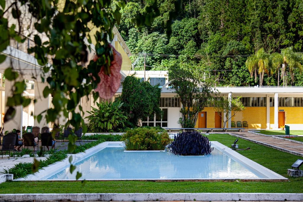 Pátio do Balneário de Águas de Lindóia com espelho d'água azul, jardim e edifício modernista de paredes amarelas ao fundo, diante da mata