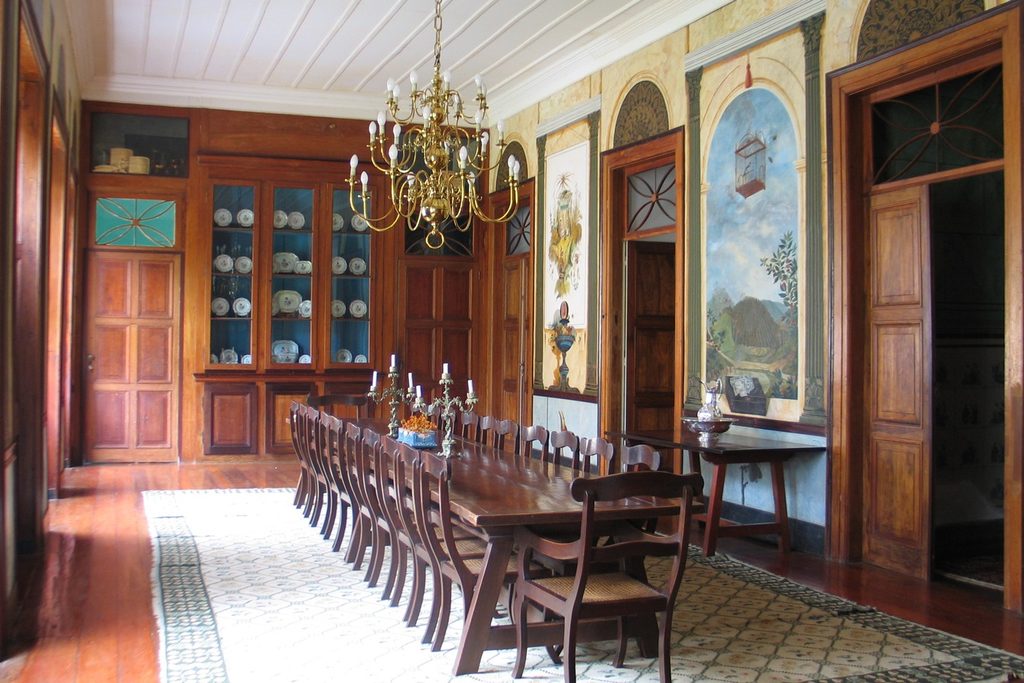 Sala de jantar com mesa longa de madeira, lustre dourado e paredes com pinturas de paisagem e gaiola, na Fazenda Resgate