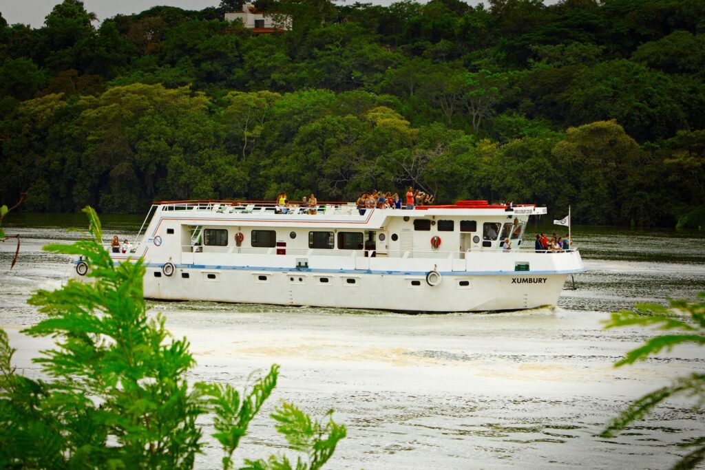Barco de turismo branco com passageiros no convés navegando pelo rio Tietê, com mata densa na margem