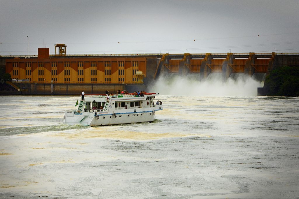 Barco de turismo no rio Tietê diante da usina hidrelétrica e da eclusa de Barra Bonita, com água saindo dos vertedouros