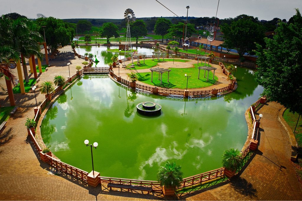 Lagoa verde com fonte no centro, cercada por passarela e gradil de madeira, em praça arborizada com moinho de vento ao fundo