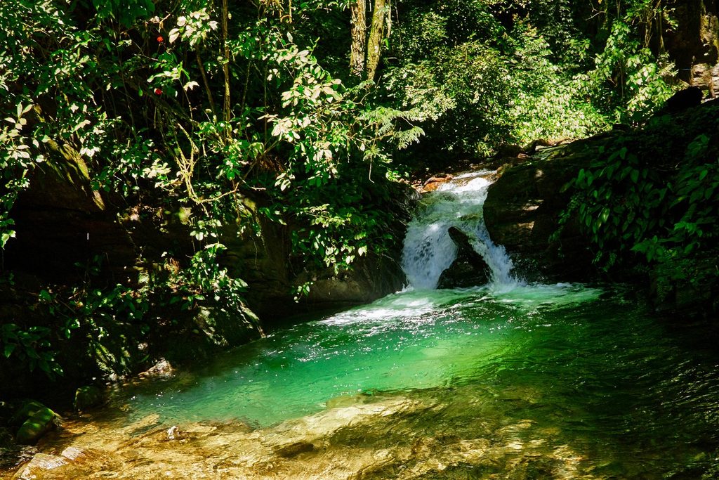 Queda d'água entre pedras e mata descendo para um poço de água verde-esmeralda