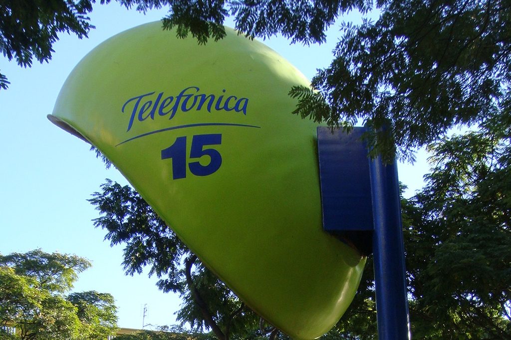 Orelhão gigante roxo sobre um poste, entre árvores e postes de luz redondos, com casarões coloridos ao fundo e pessoas passando, em Itu