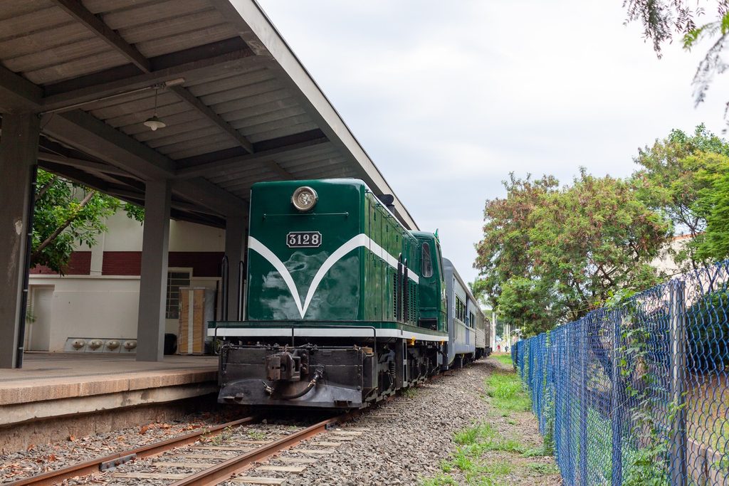 Locomotiva diesel verde e branca com o número 3128 ao lado de uma plataforma coberta, com vagões atrás e árvores ao fundo