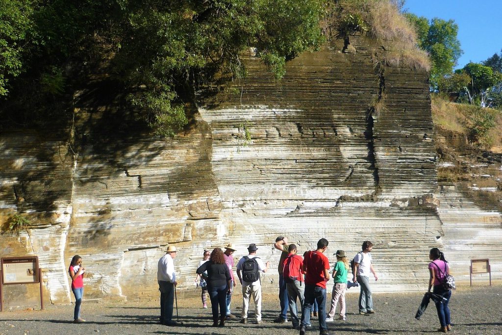 Grupo de visitantes diante de um paredão de rocha em camadas finas horizontais, no Parque do Varvito