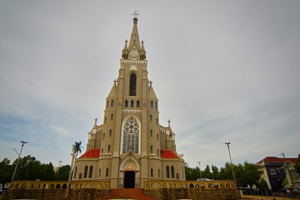 Igreja neogótica de torre alta e pontiaguda com relógio e grande rosácea na fachada, sob céu nublado