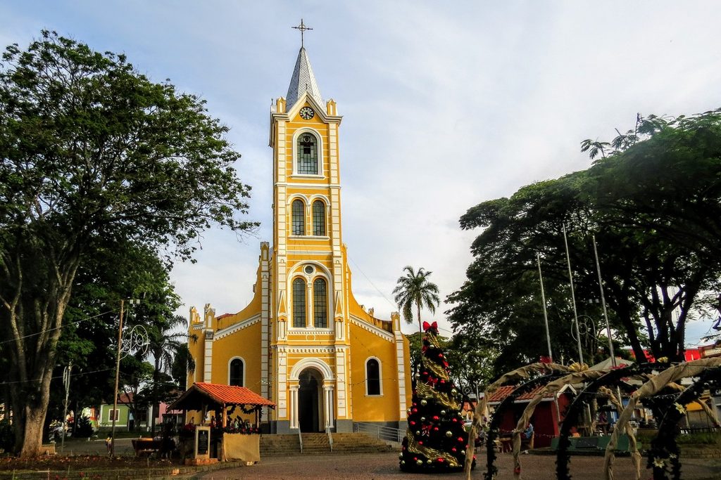Igreja matriz amarela de torre alta na praça central de Joanópolis, com árvores e decoração de Natal