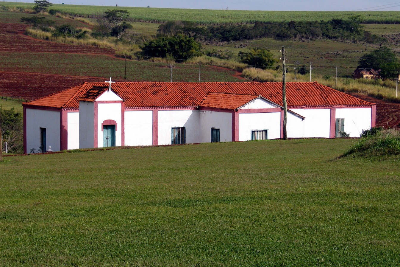 Construção branca de telhado vermelho com pequena capela e cruz na Fazenda Mandaguahy, diante de um gramado e de lavouras
