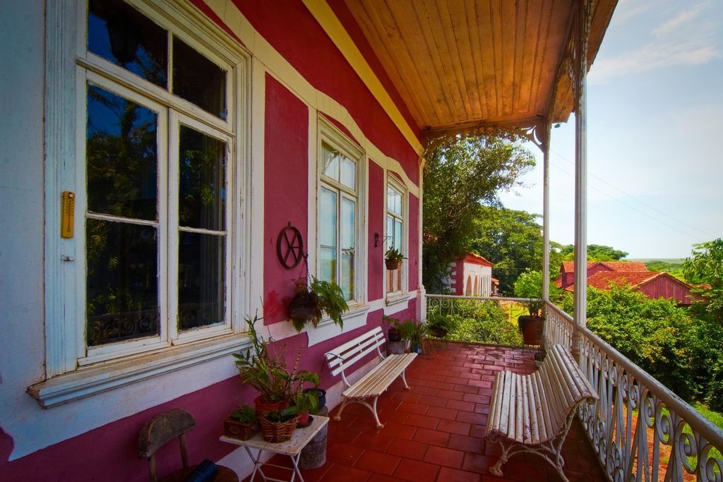 Varanda da casa-grande com paredes vermelhas, janelas brancas, bancos de ferro e vista para o pomar e telhados da fazenda