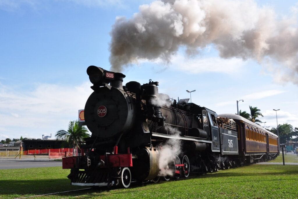 Locomotiva a vapor 505 da Maria Fumaça soltando uma nuvem de fumaça branca, puxando vagões de madeira sob céu azul