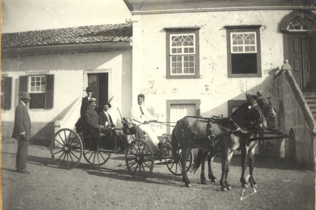Fotografia antiga em preto e branco da sede da fazenda, com carruagem puxada por cavalos e homens de chapéu na frente