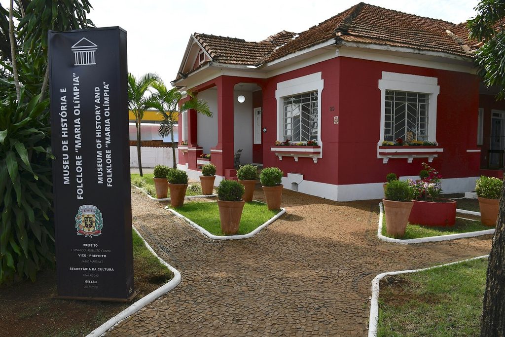 Casa térrea vermelha com janelas brancas e jardim na frente, com placa do Museu de História e Folclore Maria Olímpia