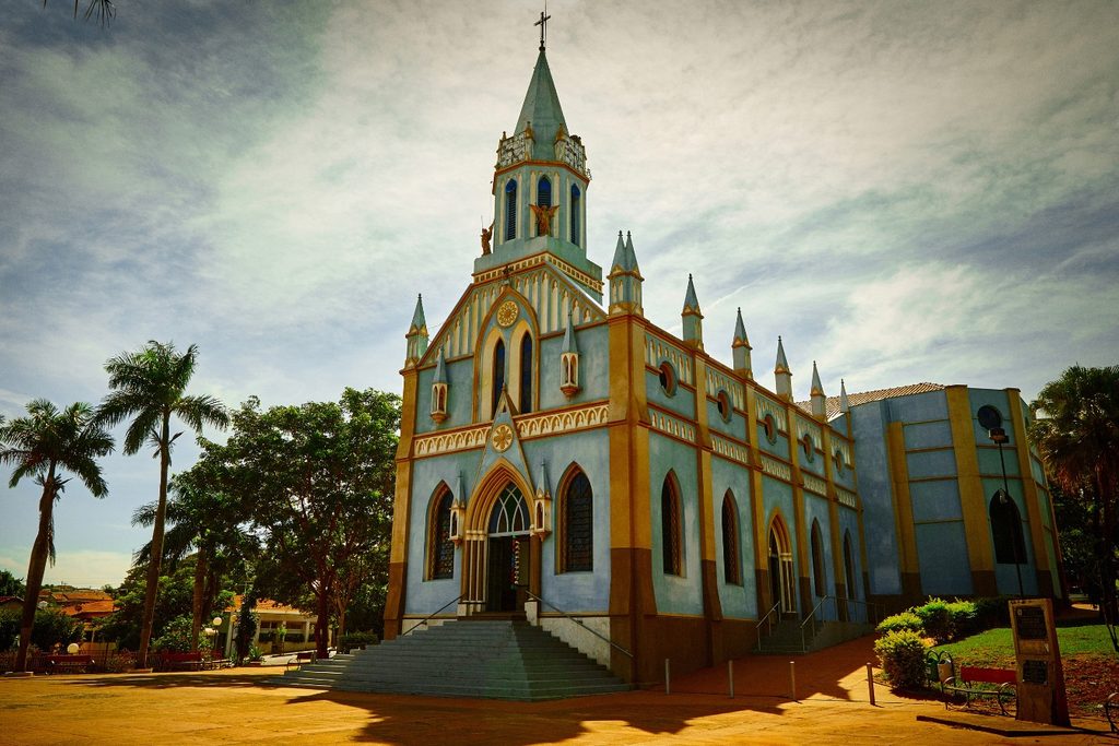 Igreja de torres pontiagudas pintada de azul e amarelo, com escadaria e palmeiras, o Santuário de Nossa Senhora Aparecida, em Olímpia