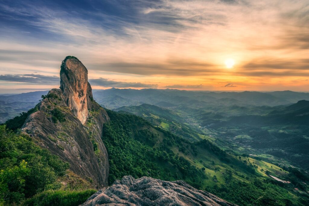 Paredão alto e estreito da Pedra do Baú ao pôr do sol, sobre montanhas verdes da Serra da Mantiqueira