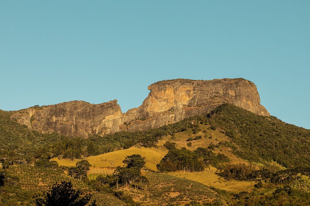 Formações rochosas do Complexo da Pedra do Baú vistas de longe, sobre morros de pasto e mata, com céu azul