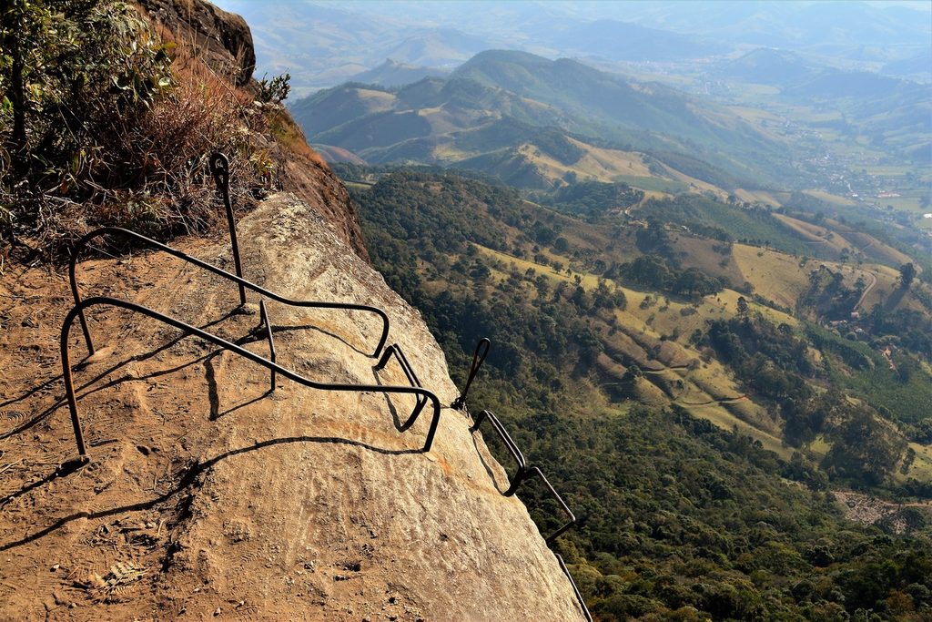 Degraus e corrimãos de ferro cravados na rocha da Pedra do Baú, com vale e montanhas ao fundo