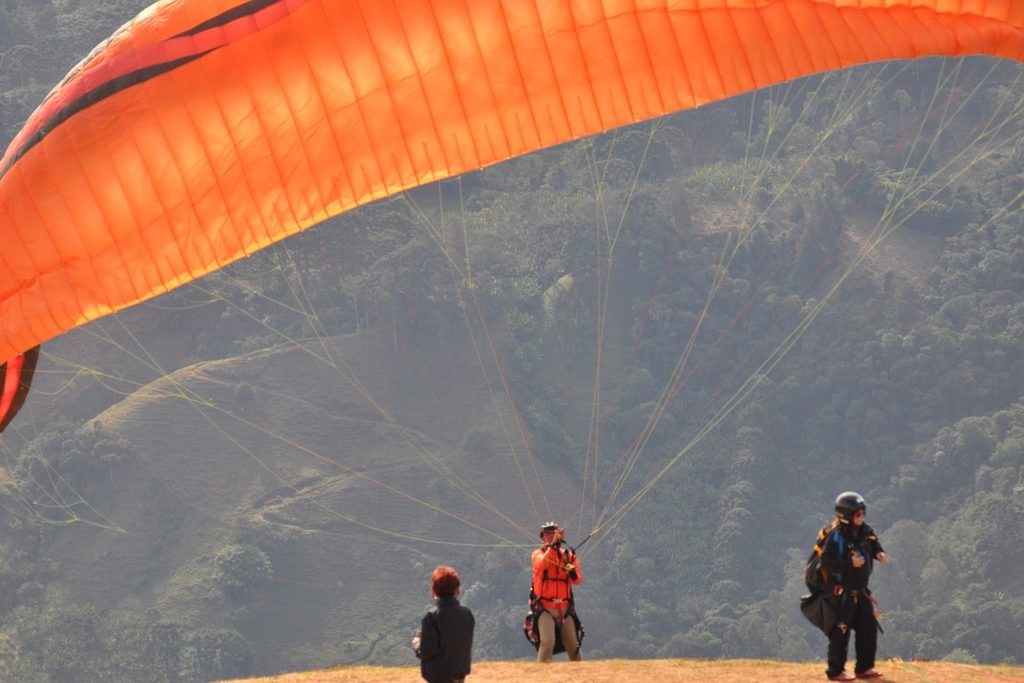 Pilotos de parapente se preparando para decolar com a vela laranja aberta, na rampa sobre o vale