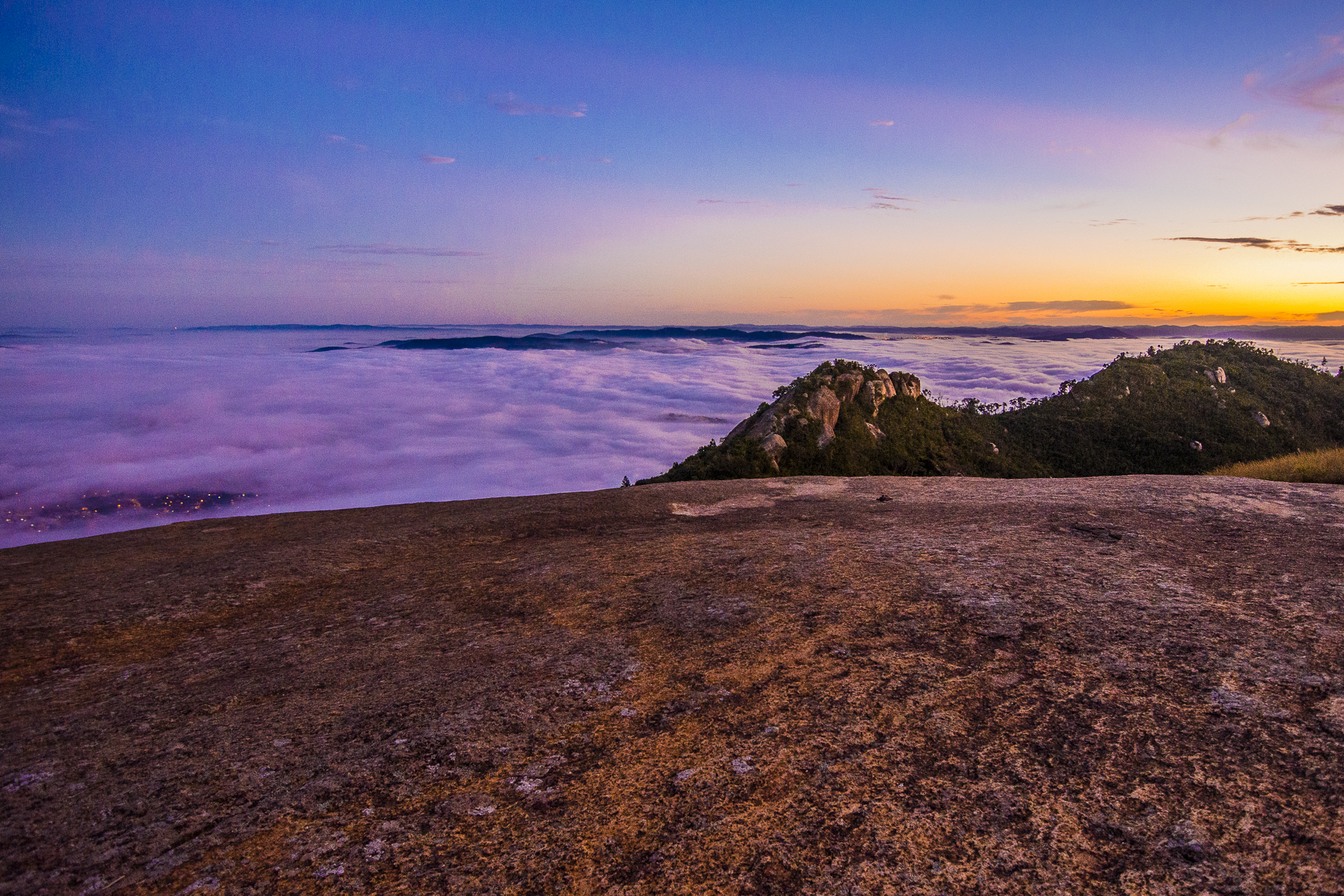 Laje de granito no topo da Pedra Grande e mar de nuvens sob um céu roxo e laranja