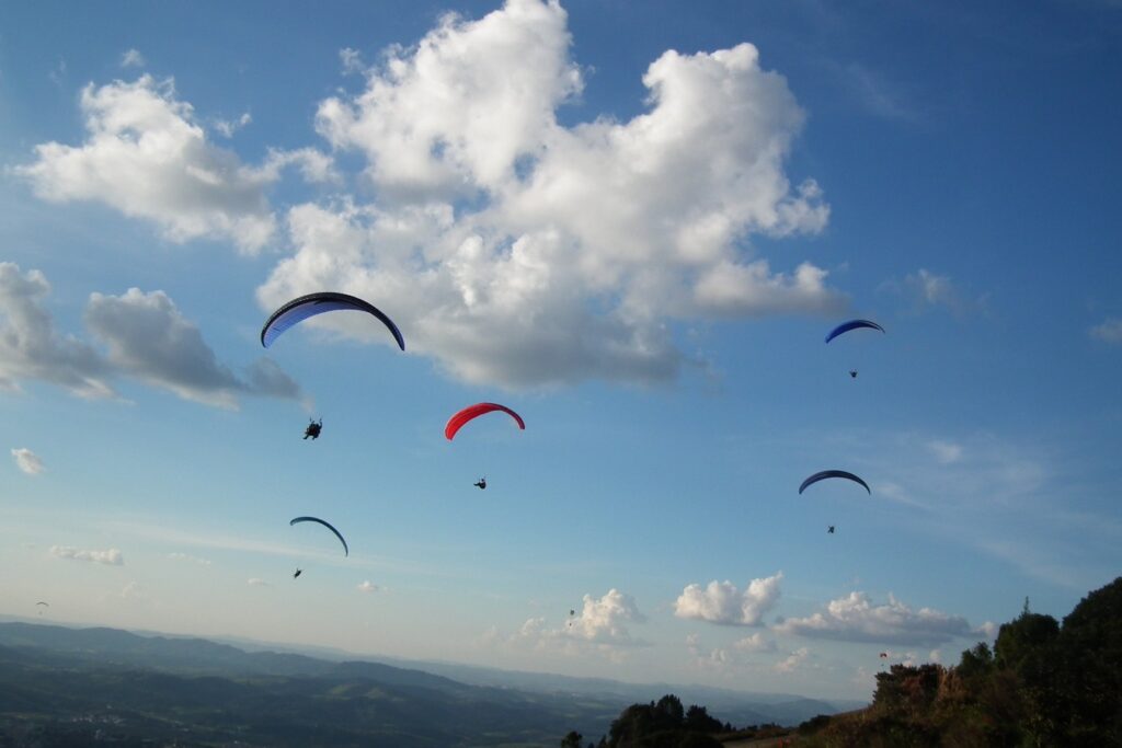 Parapentes coloridos voando sobre a Pedra Grande, com nuvens e serras ao fundo