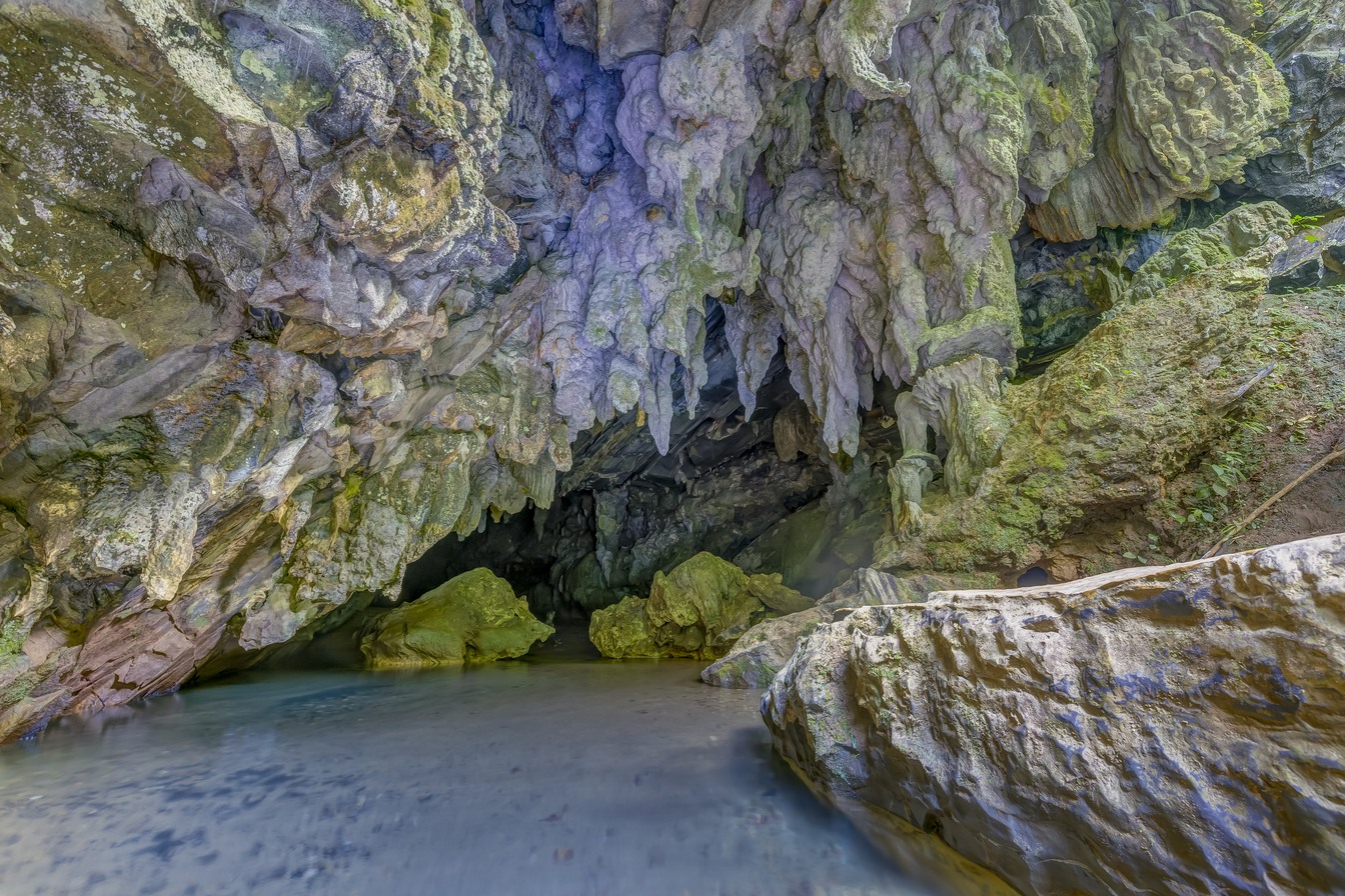 Entrada de uma caverna do PETAR, com estalactites e paredes de pedra cobertas de musgo sobre uma poça de água azulada