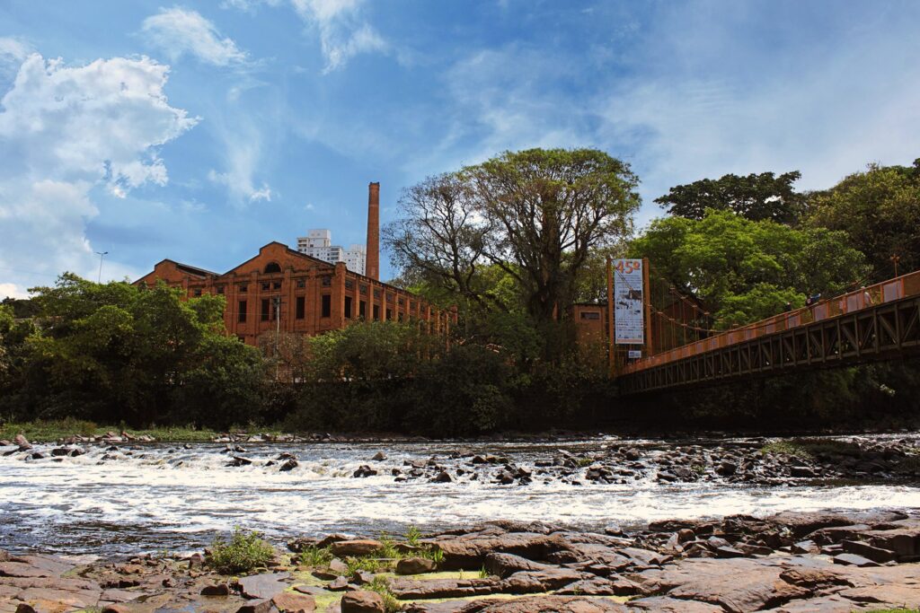 Corredeiras do rio Piracicaba sobre pedras, com os prédios de tijolo do Engenho Central e a passarela pênsil ao fundo