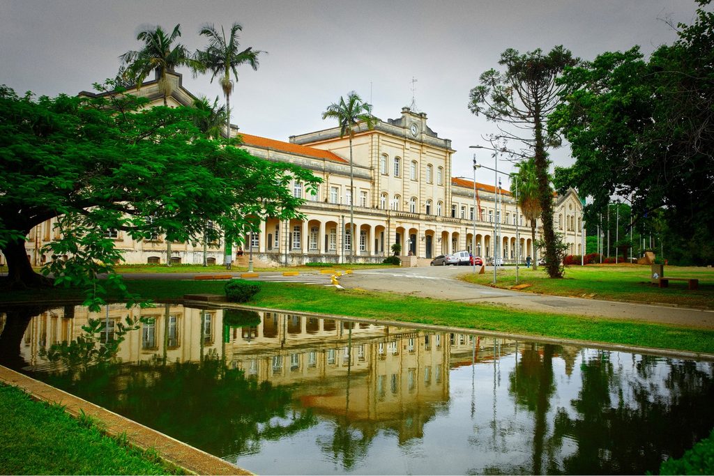 Prédio central amarelo da Esalq refletido num espelho d'água entre gramados, palmeiras e árvores