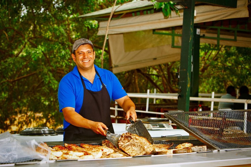 Cozinheiro sorridente de avental servindo peixes assados numa grelha grande em restaurante da Rua do Porto