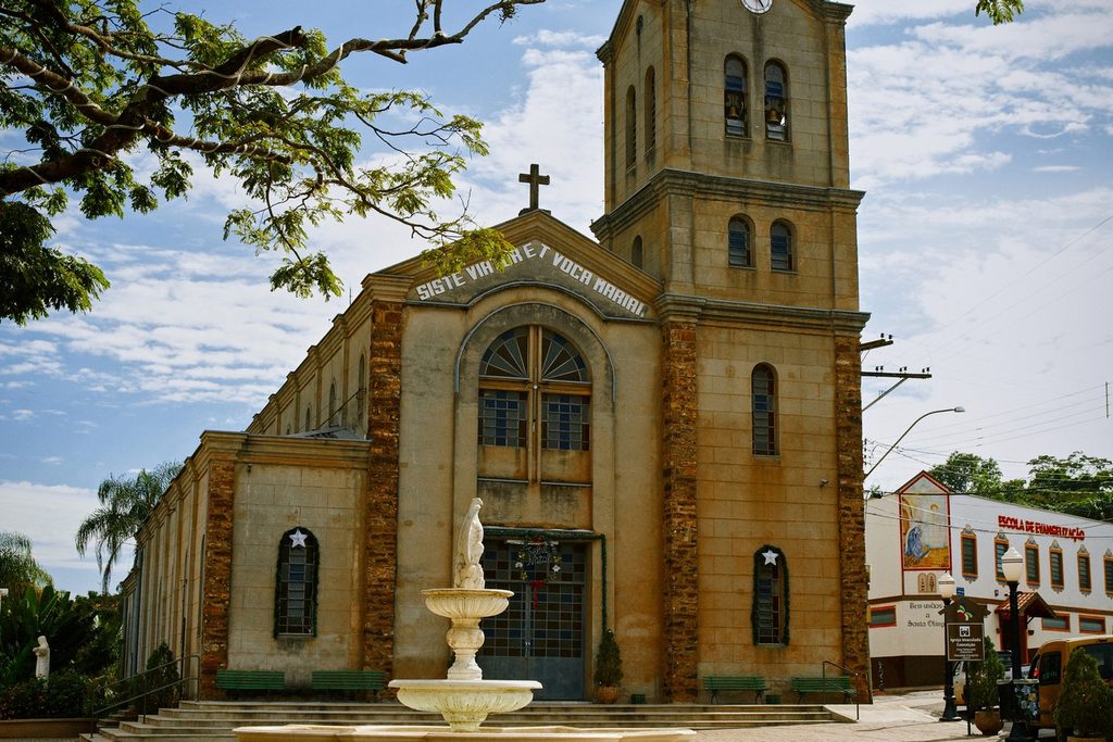 Igreja de pedra com torre do relógio e chafariz na frente, num dos bairros tiroleses de Santana e Santa Olímpia