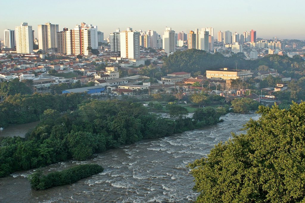 Vista do alto do rio Piracicaba com corredeiras e mata nas margens, e prédios da cidade ao fundo
