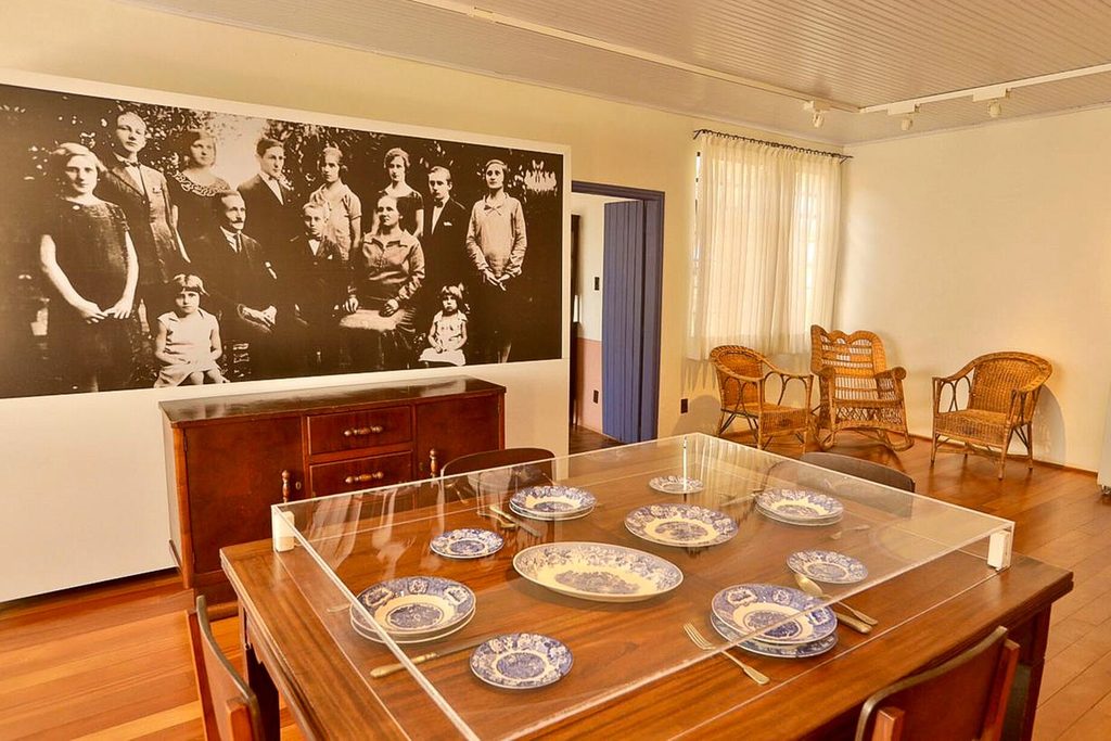 Sala de jantar com mesa, louças antigas protegidas por vidro e grande fotografia da família Portinari na parede