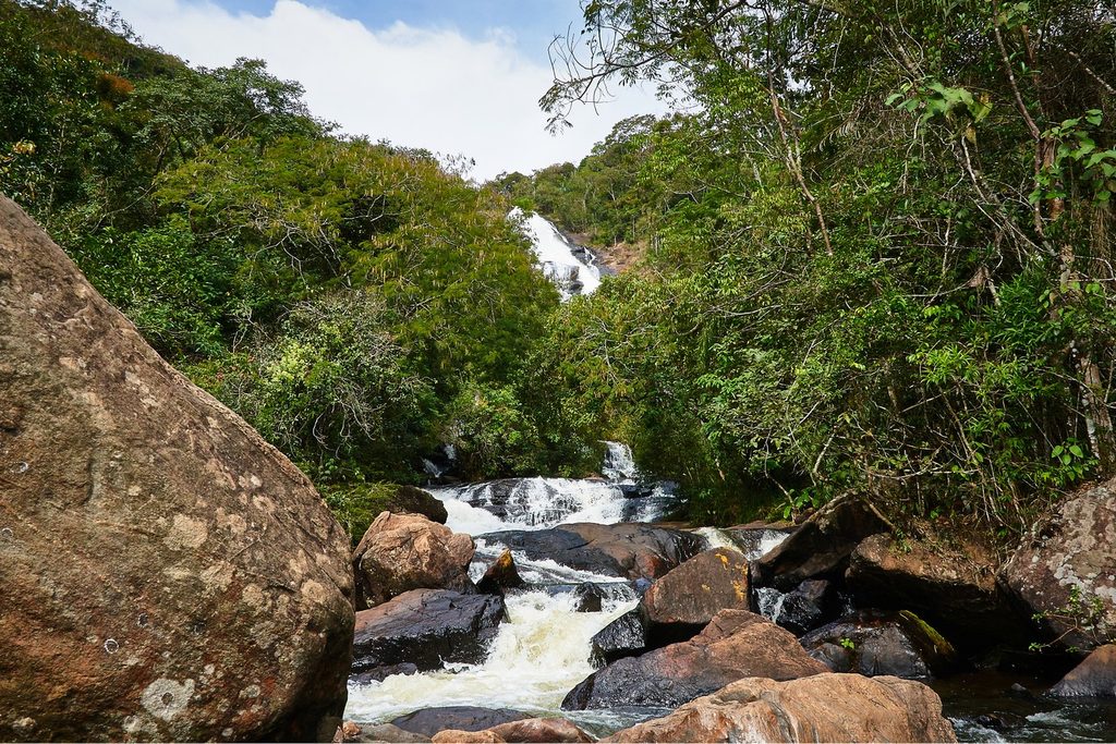Rio correndo entre grandes pedras no meio da mata, com a queda da Cachoeira dos Pretos ao fundo