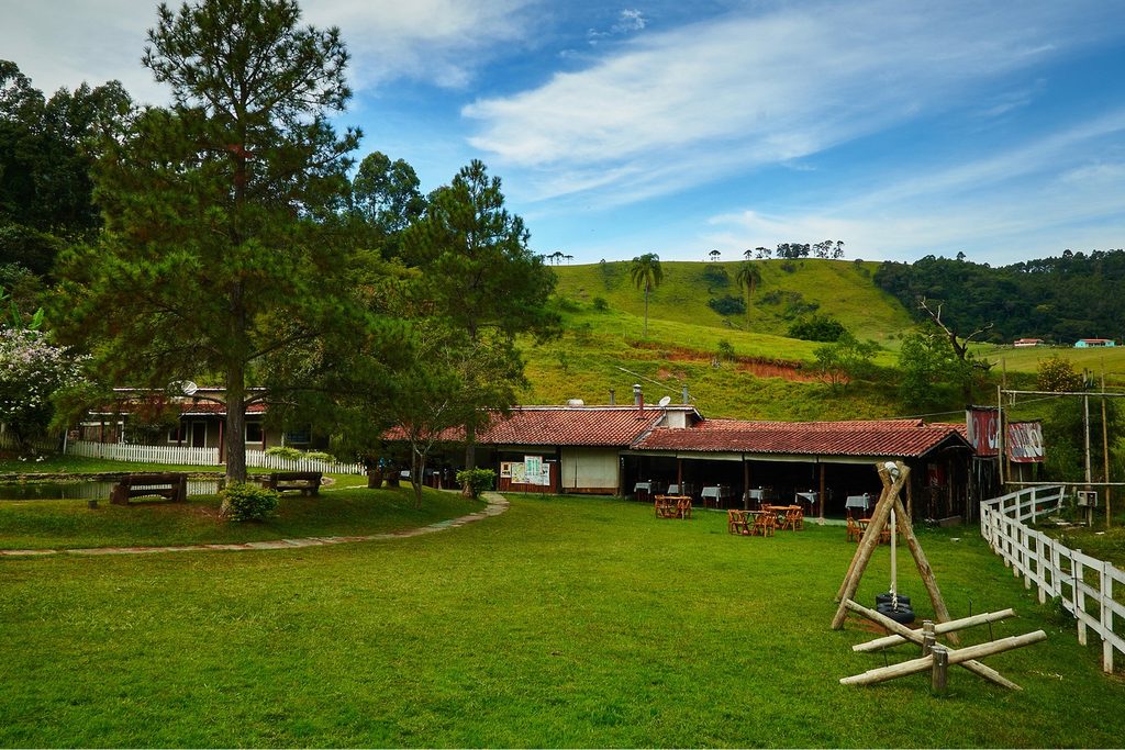 Restaurante rural de telhado vermelho com mesas ao ar livre num gramado cercado de pinheiros e morros verdes