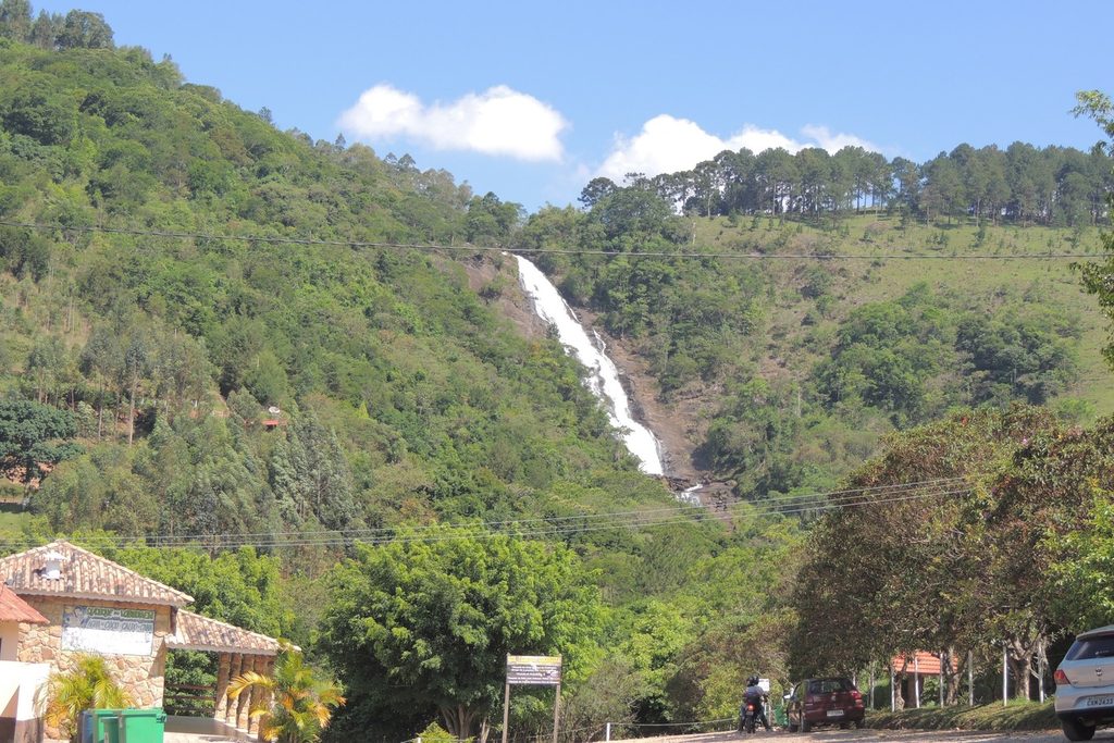 Queda longa e branca da Cachoeira dos Pretos na encosta verde, com construções e carros na estrada em primeiro plano