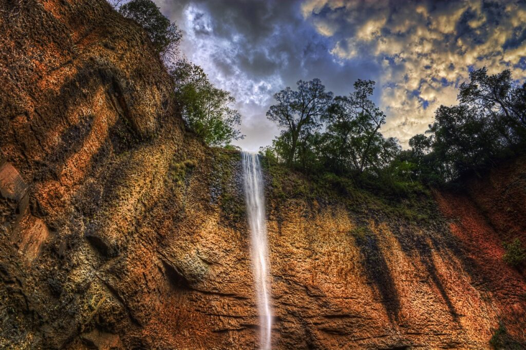 Cachoeira estreita e alta despencando por um paredão de rocha alaranjada, com árvores no alto e céu de nuvens