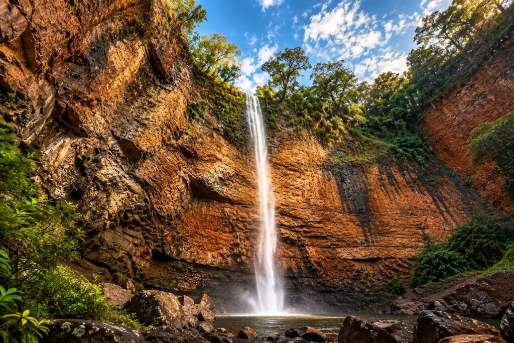 Cachoeira alta e estreita caindo por um paredão de rocha alaranjada até um poço, cercada de mata e sob céu azul