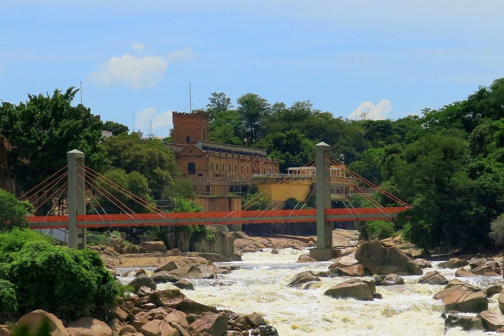 Ponte pênsil vermelha sobre as corredeiras do Rio Tietê, com um prédio antigo de tijolos ao fundo, em Salto