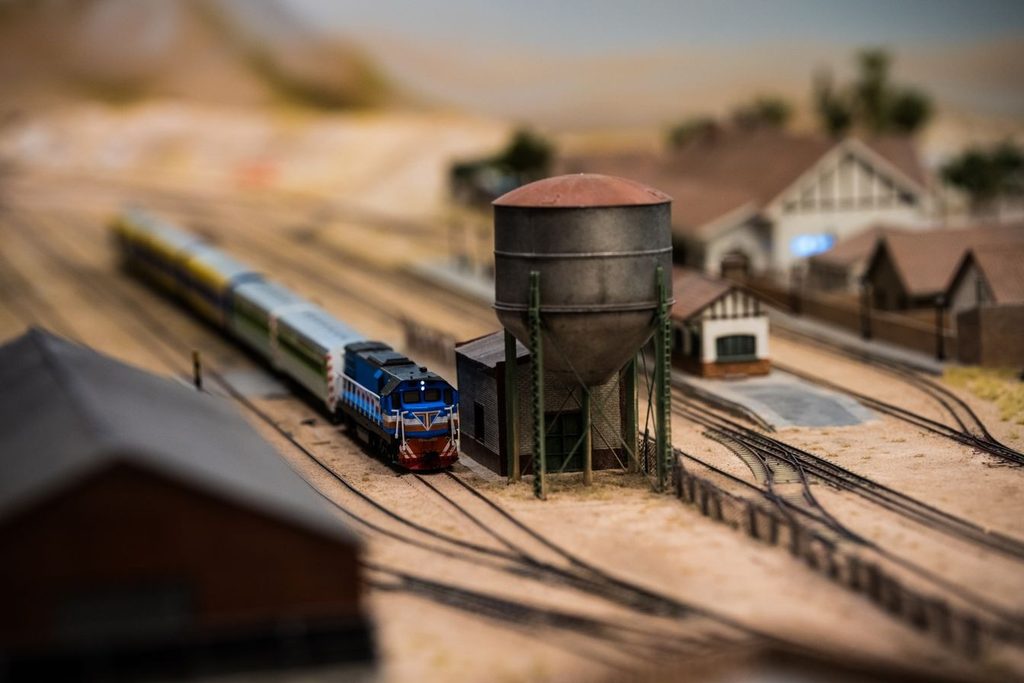 Maquete ferroviária em miniatura com trem azul, caixa d'água e casas de estação, em imagem ilustrativa
