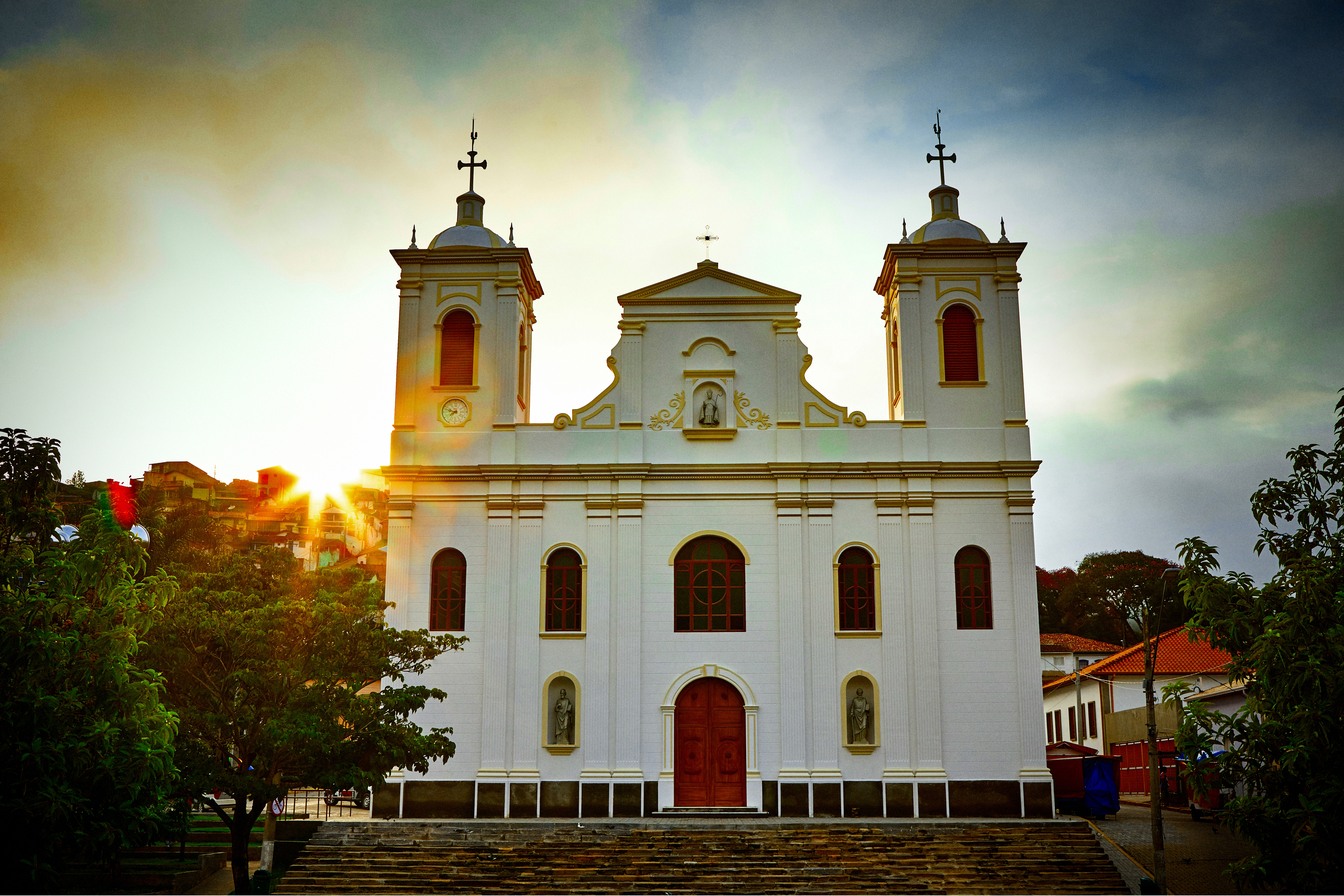 Igreja Matriz branca de duas torres de São Luiz do Paraitinga no fim de tarde, com o sol surgindo atrás das árvores
