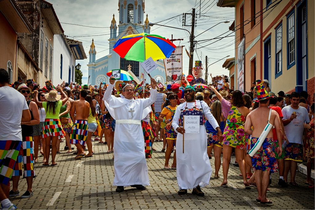 Foliões fantasiados e guarda-chuva colorido em bloco de carnaval na rua de São Luiz do Paraitinga, com a Igreja Matriz ao fundo