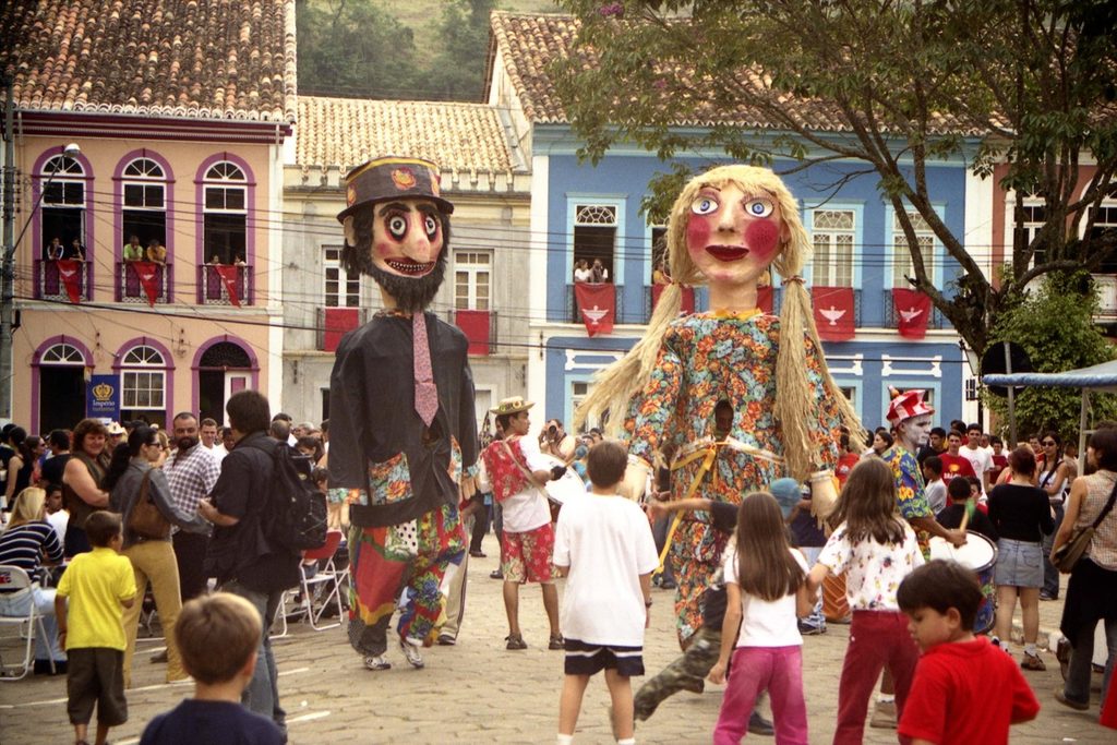 Bonecões gigantes João Paulino e Maria Angu dançando na praça da Festa do Divino, entre crianças e casarões coloridos
