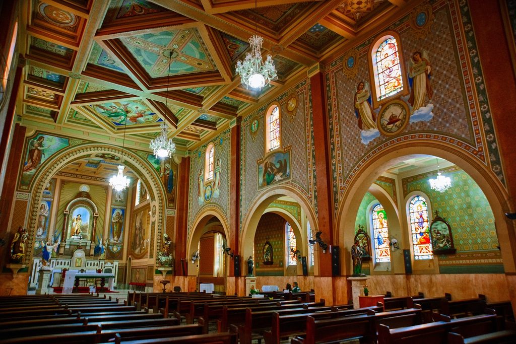 Interior da Igreja Matriz de São Roque, com teto e paredes pintados, arcos, vitrais coloridos, lustres e bancos de madeira