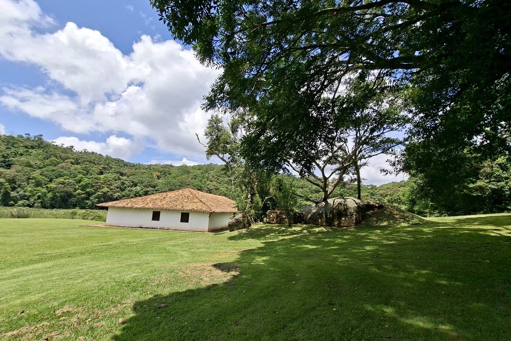 Casa grande térrea de paredes brancas e telhado de telhas, em gramado amplo sob árvores, com morros de mata ao fundo, no Sítio Santo Antônio