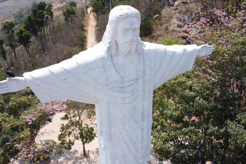 Estátua branca do Cristo Redentor de braços abertos, vista de cima, entre a vegetação do mirante