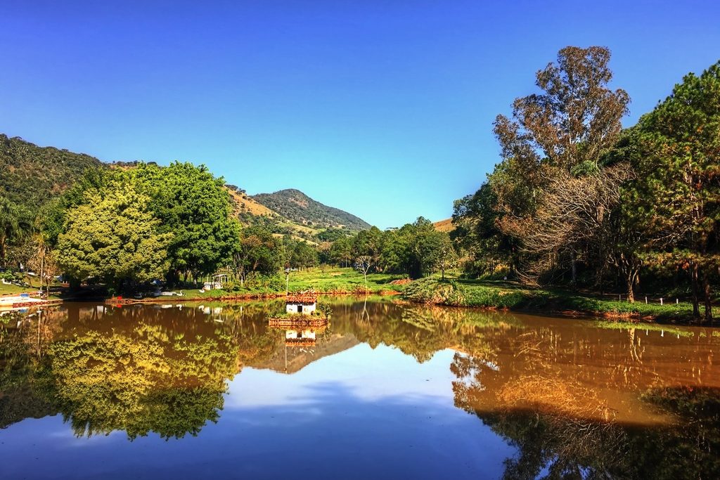 Lago de águas calmas refletindo árvores e morros sob céu azul na zona rural de Socorro, SP