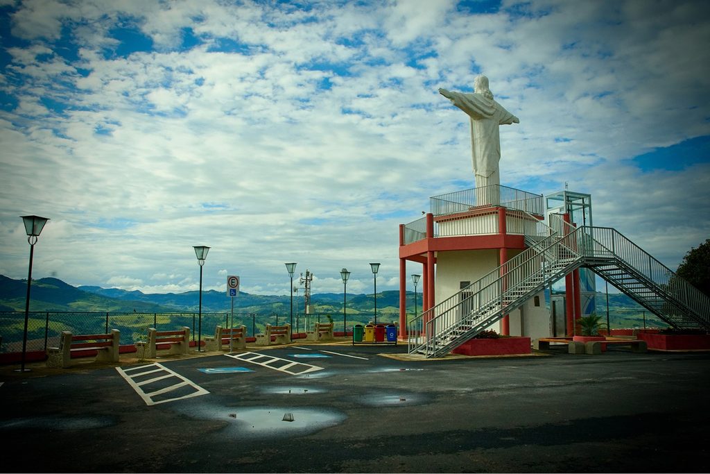 Estátua branca do Cristo sobre base vermelha no Mirante do Cristo, com montanhas ao fundo e céu com nuvens