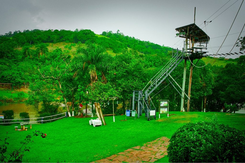 Torre de tirolesa com escada metálica num gramado verde cercado de mata, no Parque do Monjolinho, em Socorro