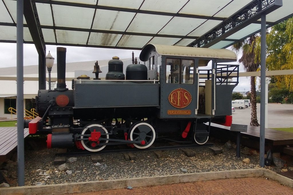 Pequena locomotiva a vapor preta com rodas vermelhas e símbolo da Sorocabana, exposta sob uma cobertura
