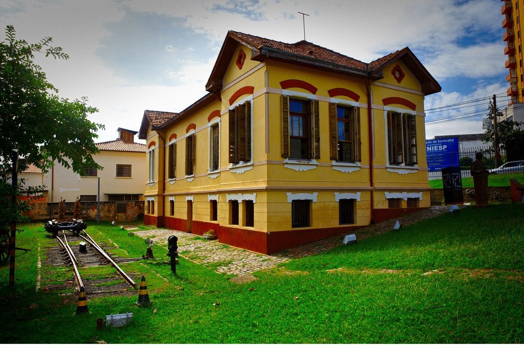 Casarão amarelo com trilhos e peças ferroviárias no gramado, o Museu da Estrada de Ferro Sorocabana, bom complemento ao passeio do Trem dos Operários