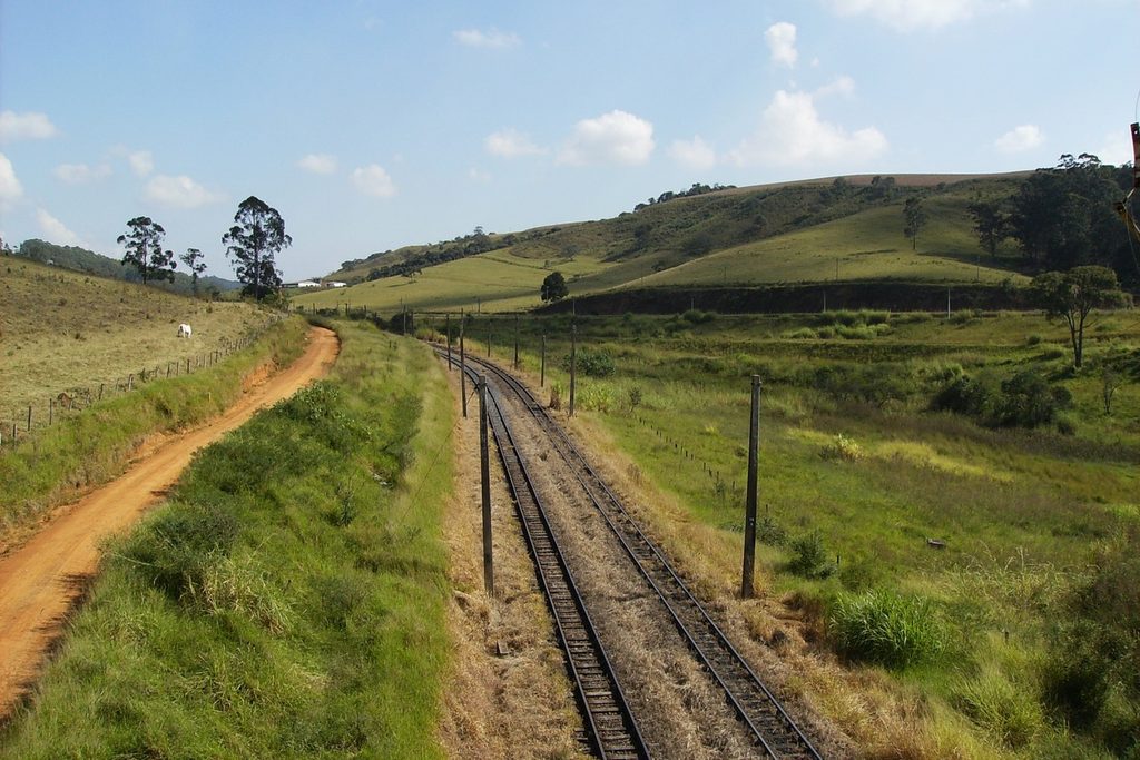 Trilhos duplos da antiga Estrada de Ferro Sorocabana fazendo curva entre pastos verdes e morros, ao lado de uma estrada de terra