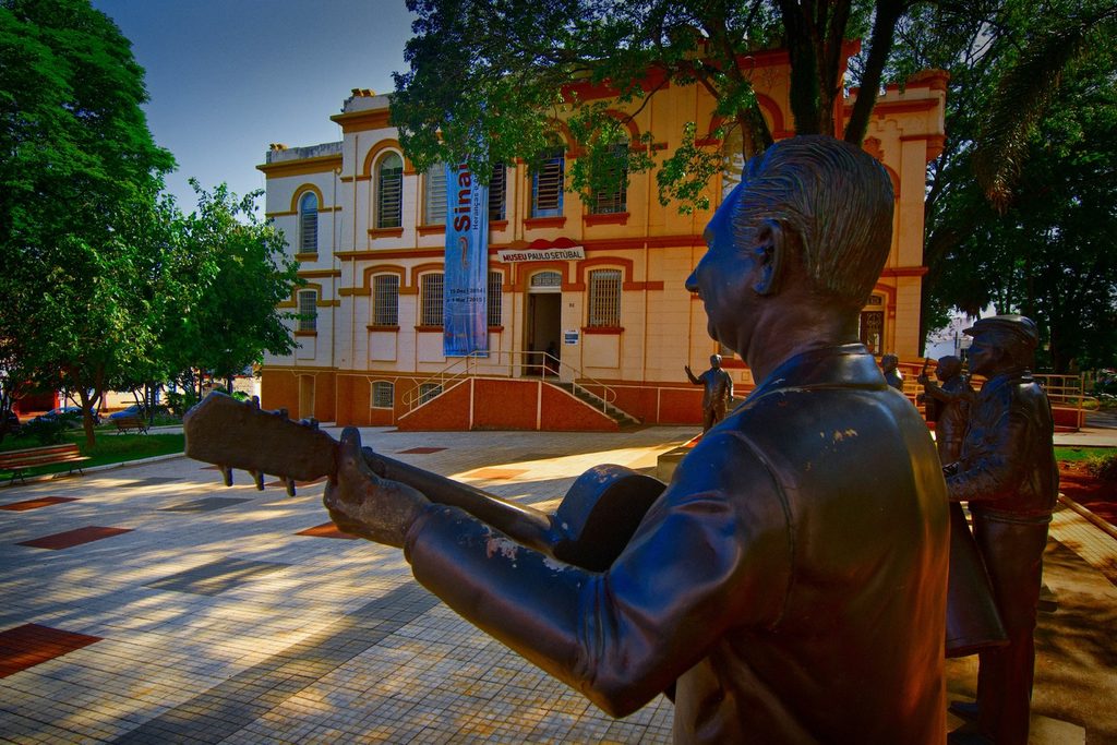 Estátua de bronze de um violeiro em primeiro plano diante do prédio amarelo e vermelho do Museu Histórico Paulo Setúbal